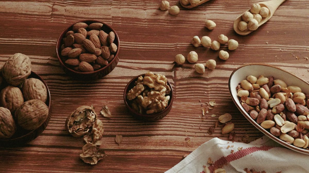 A top view of various nuts in bowls and spoons on a wooden tabletop.